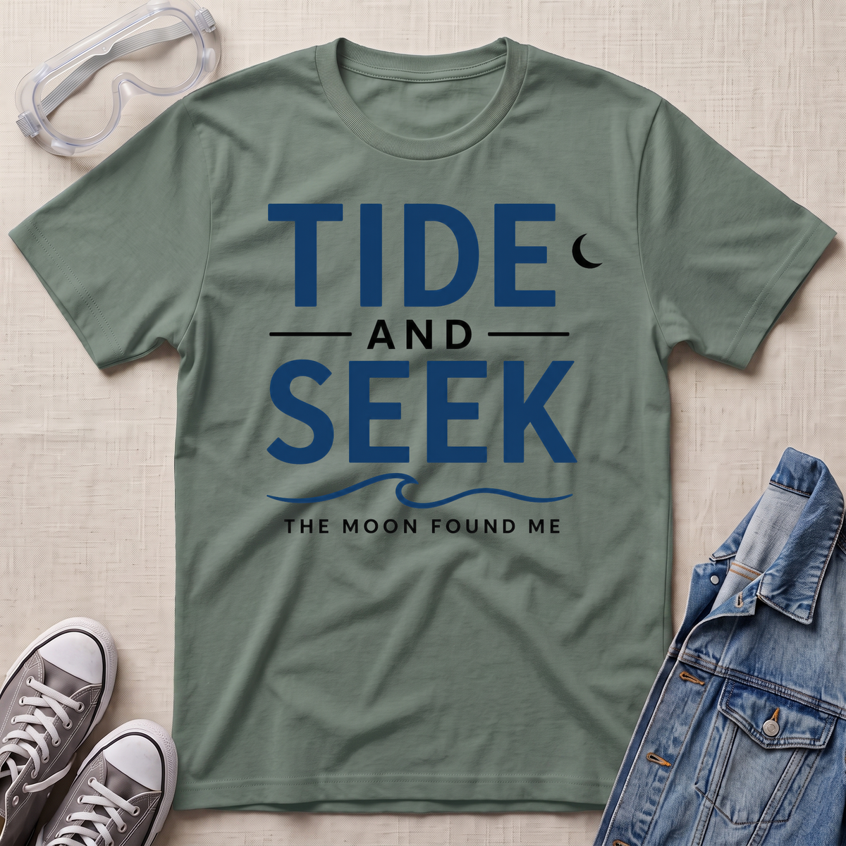 A sage green short-sleeve graphic T-shirt laid flat on a light beige textured surface, with safety glasses, gray sneakers, and a folded denim jacket around it. The shirt has large navy text reading 'TIDE AND SEEK' with smaller text 'THE MOON FOUND ME' and a crescent moon graphic.