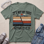 An olive green graphic t-shirt laid flat on a textured beige background, styled with safety glasses, gray sneakers, and a blue denim jacket. The t-shirt features a retro-style cross-section graphic of earth layers with a crack running through the center, accompanied by the text 'IT'S NOT MY FAULT ACTUALLY, IT IS'.