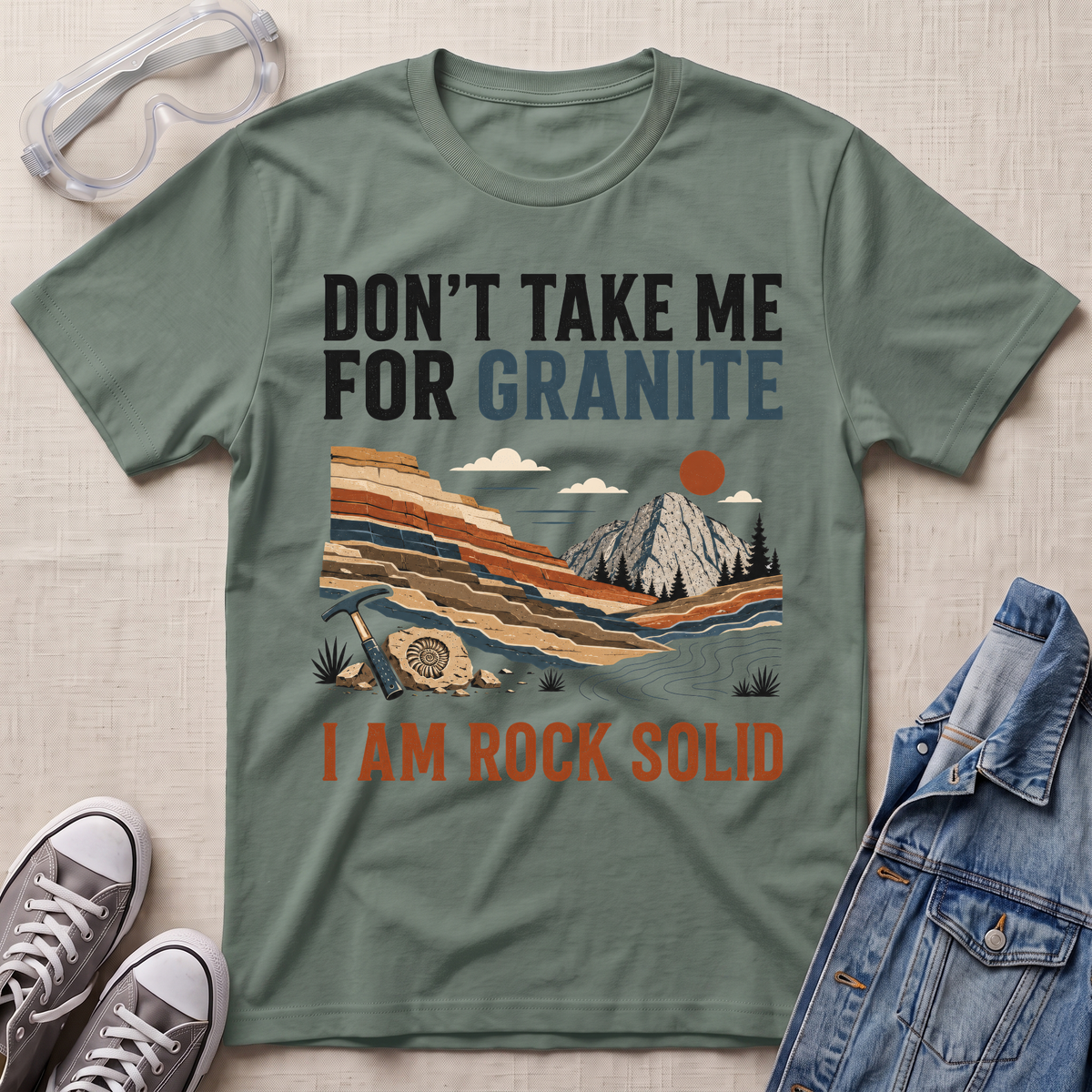 A sage green graphic t-shirt laid flat on a textured beige fabric background, styled with safety goggles, gray sneakers, and a blue denim jacket. The t-shirt features a graphic of a mountain landscape with the text 'DON'T TAKE ME FOR GRANITE' and 'I AM ROCK SOLID'.