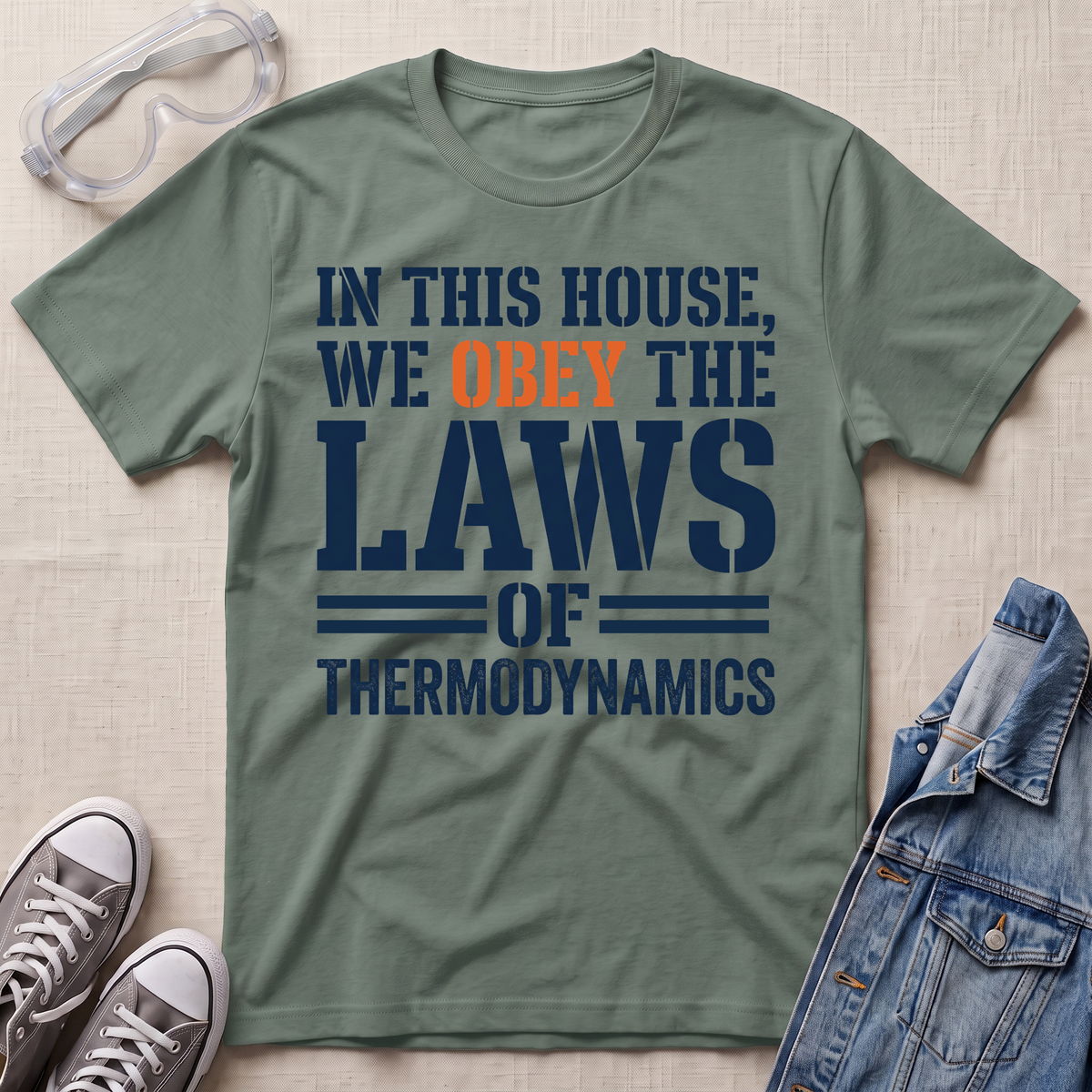An olive green graphic t-shirt laid flat on a beige fabric background, styled with safety goggles, gray sneakers, and a denim jacket. The t-shirt features the text 'IN THIS HOUSE, WE OBEY THE LAWS OF THERMODYNAMICS' in navy blue and orange.