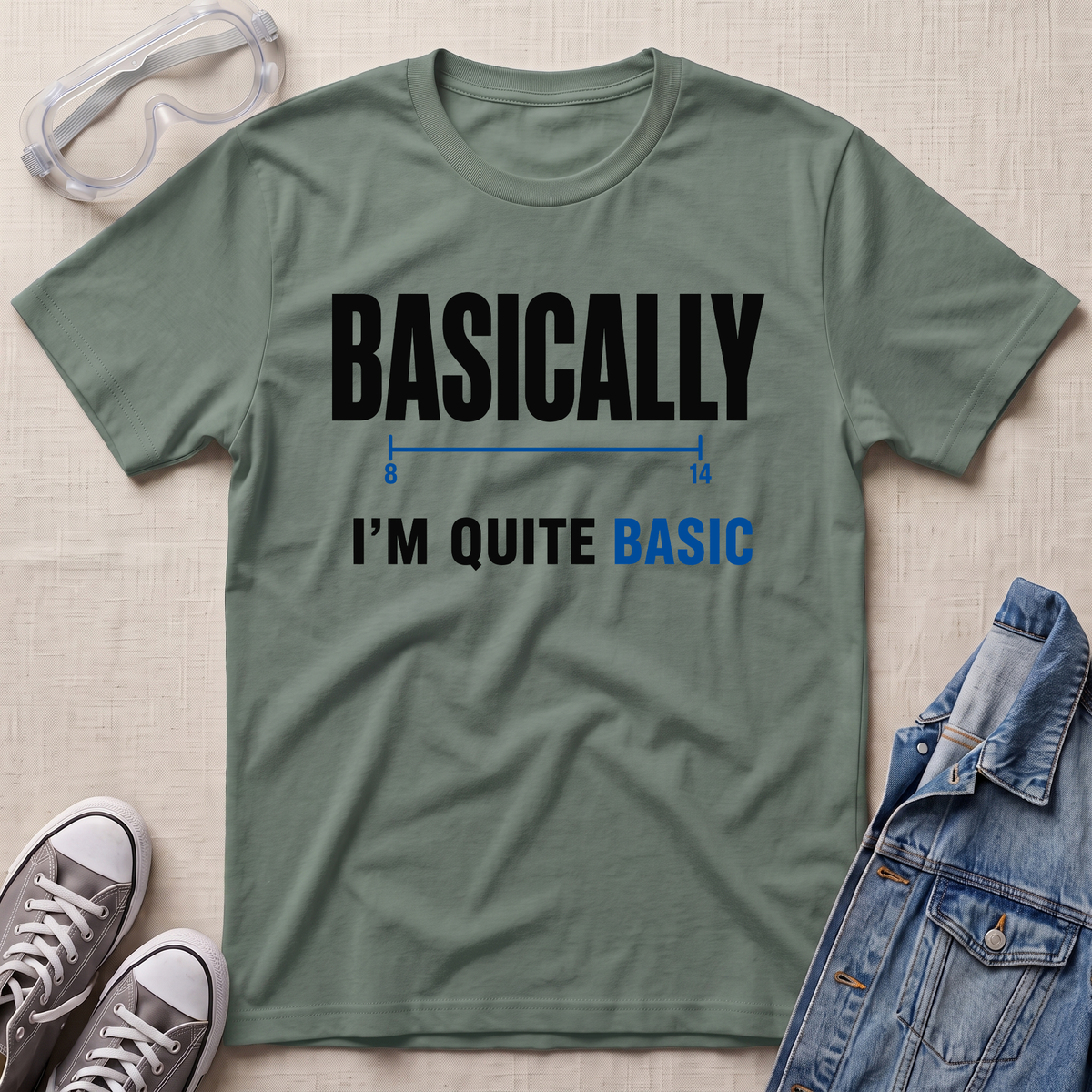 An olive green graphic t-shirt laid flat on a textured beige fabric background, styled with safety goggles, gray sneakers, and a blue denim jacket. The t-shirt features the text 'BASICALLY I'M QUITE BASIC' in black and blue lettering.