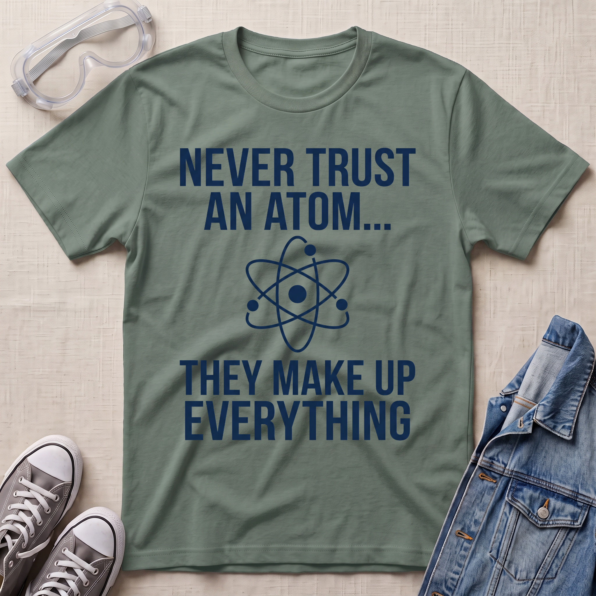 A olive green graphic t-shirt with white text reading 'NEVER TRUST AN ATOM.' and 'THEY MAKE UP EVERYTHING' with an image of an atom in between the texts.