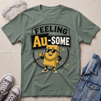 A sage green graphic t-shirt featuring a cartoon gold bar character wearing sunglasses and a gold chain with the text 'FEELING Au-SOME'. The t-shirt is styled in a flat-lay arrangement with clear safety goggles, gray sneakers, and a blue denim jacket on a textured beige background.