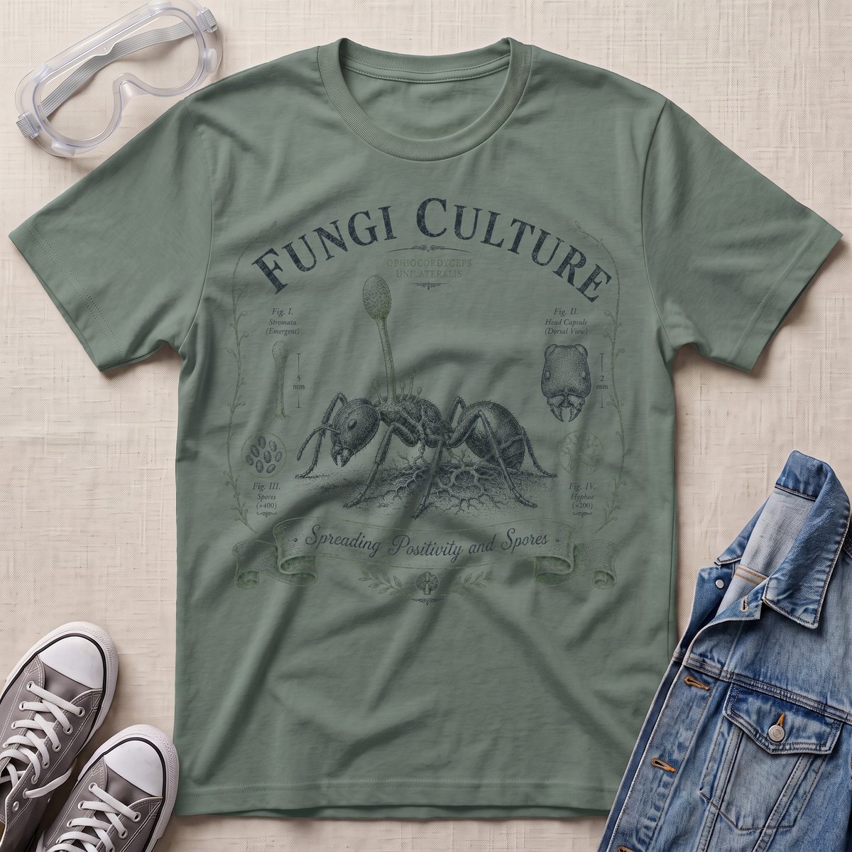 An olive green graphic t-shirt featuring a vintage-style ant illustration and the text 'Fungi Culture' and 'Spreading Butterflies and Spiders'. The t-shirt is styled in a flat-lay arrangement on a beige textured background, accompanied by safety goggles, gray sneakers, and a blue denim jacket.