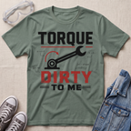 A sage green graphic t-shirt laid flat on a beige fabric background, styled with safety glasses, gray sneakers, and a denim jacket. The t-shirt features a wrench and gear graphic with the text 'TORQUE DIRTY TO ME' in white and red.