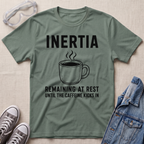 An olive green graphic t-shirt laid flat on a textured beige fabric background, surrounded by lifestyle props including safety goggles, gray sneakers, and a blue denim jacket. The t-shirt features a black graphic of a steaming coffee mug with the text 'INERTIA REMAINING AT REST UNTIL THE CAFFEINE KICKS IN'.