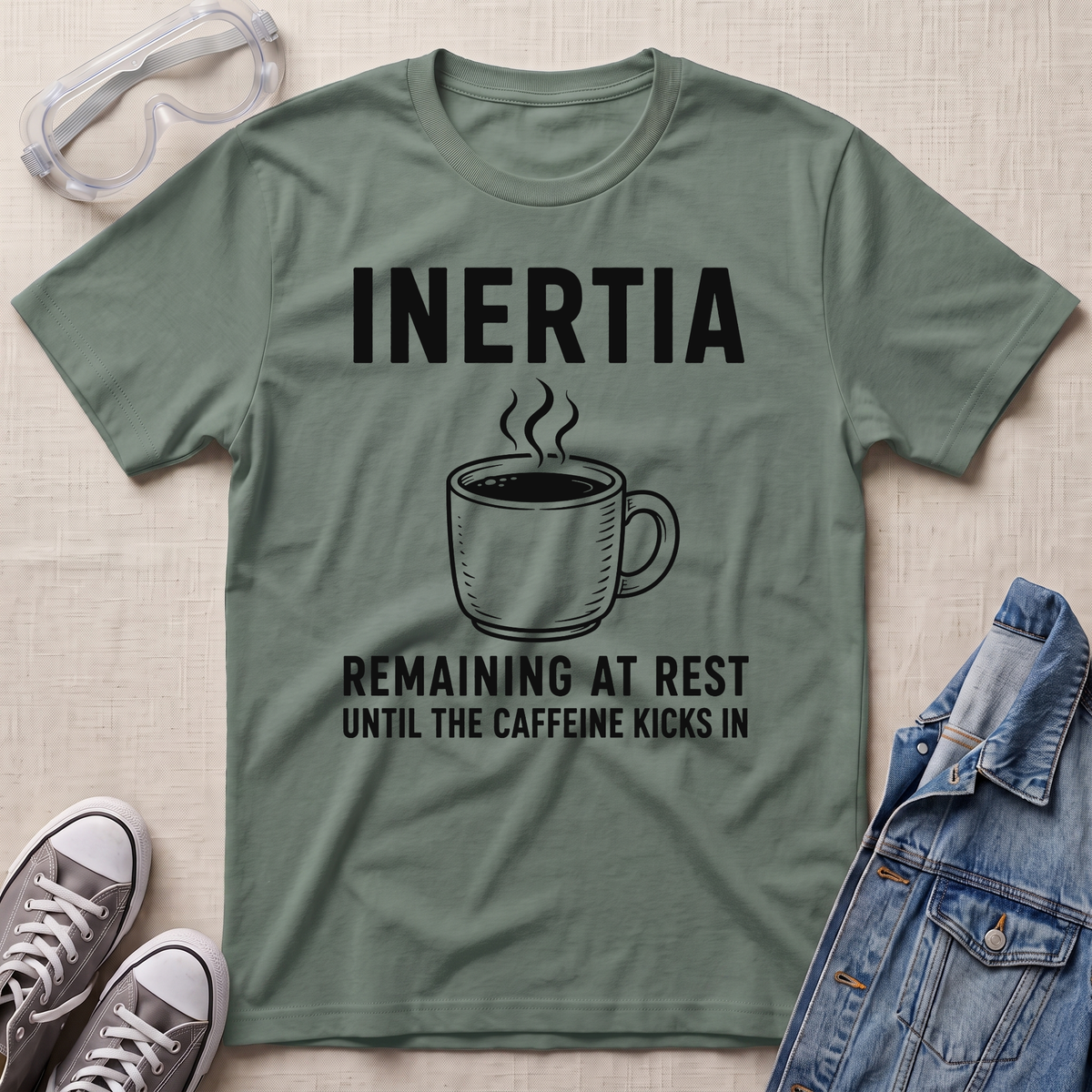 An olive green graphic t-shirt laid flat on a textured beige fabric background, surrounded by lifestyle props including safety goggles, gray sneakers, and a blue denim jacket. The t-shirt features a black graphic of a steaming coffee mug with the text 'INERTIA REMAINING AT REST UNTIL THE CAFFEINE KICKS IN'.