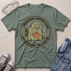 An olive green graphic t-shirt laid flat on a textured beige fabric background, surrounded by lifestyle props including safety goggles, gray sneakers, and a blue denim jacket. The t-shirt features a circular graphic of a cartoon creature in a gas mask holding a mug, with the text 'LABORATORY SURVIVAL CLUB' and 'UNDER PRESSURE, BUT ADAPTING'.