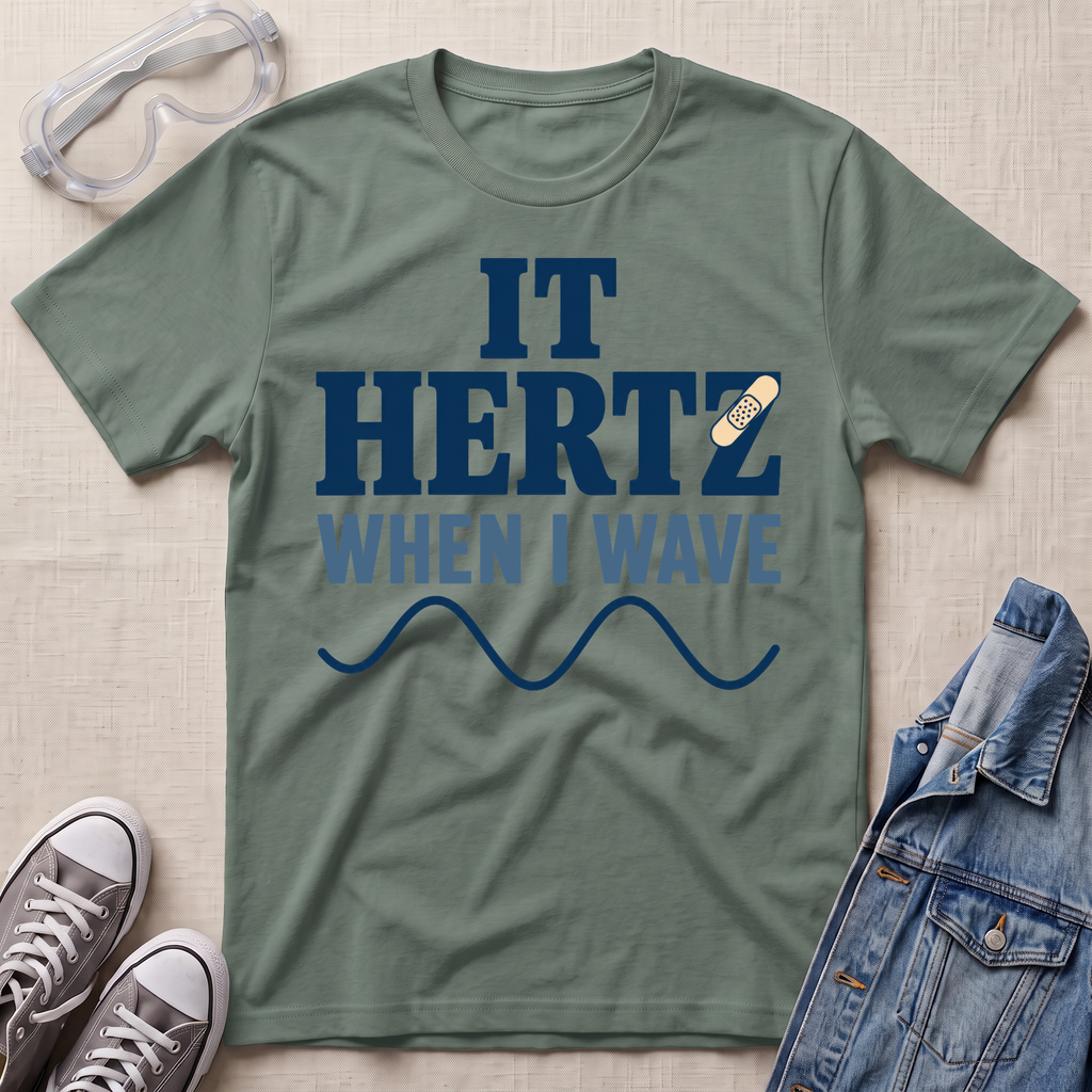 A sage green graphic t-shirt laid flat on a textured beige background, surrounded by lifestyle props including safety goggles, gray sneakers, and a denim jacket. The t-shirt features a dark blue graphic that reads 'IT HERTZ WHEN I WAVE' with a small band-aid graphic over the letter 'Z'.