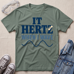 A sage green graphic t-shirt laid flat on a textured beige background, surrounded by lifestyle props including safety goggles, gray sneakers, and a denim jacket. The t-shirt features a dark blue graphic that reads 'IT HERTZ WHEN I WAVE' with a small band-aid graphic over the letter 'Z'.
