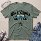 An olive green graphic t-shirt laid flat on a textured beige fabric background, styled with safety glasses, gray sneakers, and a blue denim jacket. The t-shirt features the text 'I'M NON-EUCLIDEAN BEFORE COFFEE' in black and teal lettering with a coffee cup graphic below.