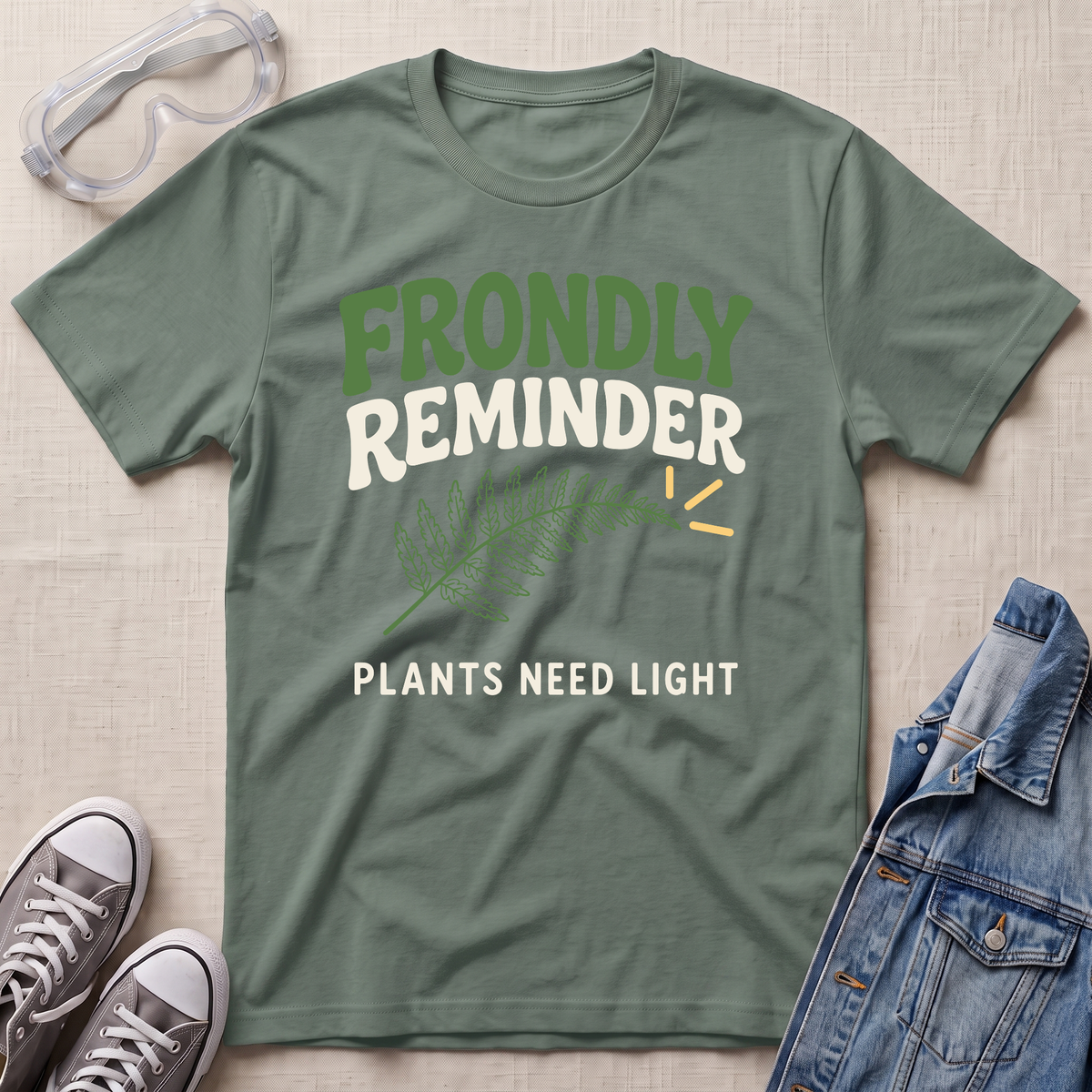 A sage green graphic t-shirt laid flat on a textured beige fabric background, surrounded by lifestyle props including clear safety goggles, gray sneakers, and a blue denim jacket. The t-shirt features a graphic that reads 'FRONDLY REMINDER' in green and white, with a green fern illustration and 'PLANTS NEED LIGHT' printed below.