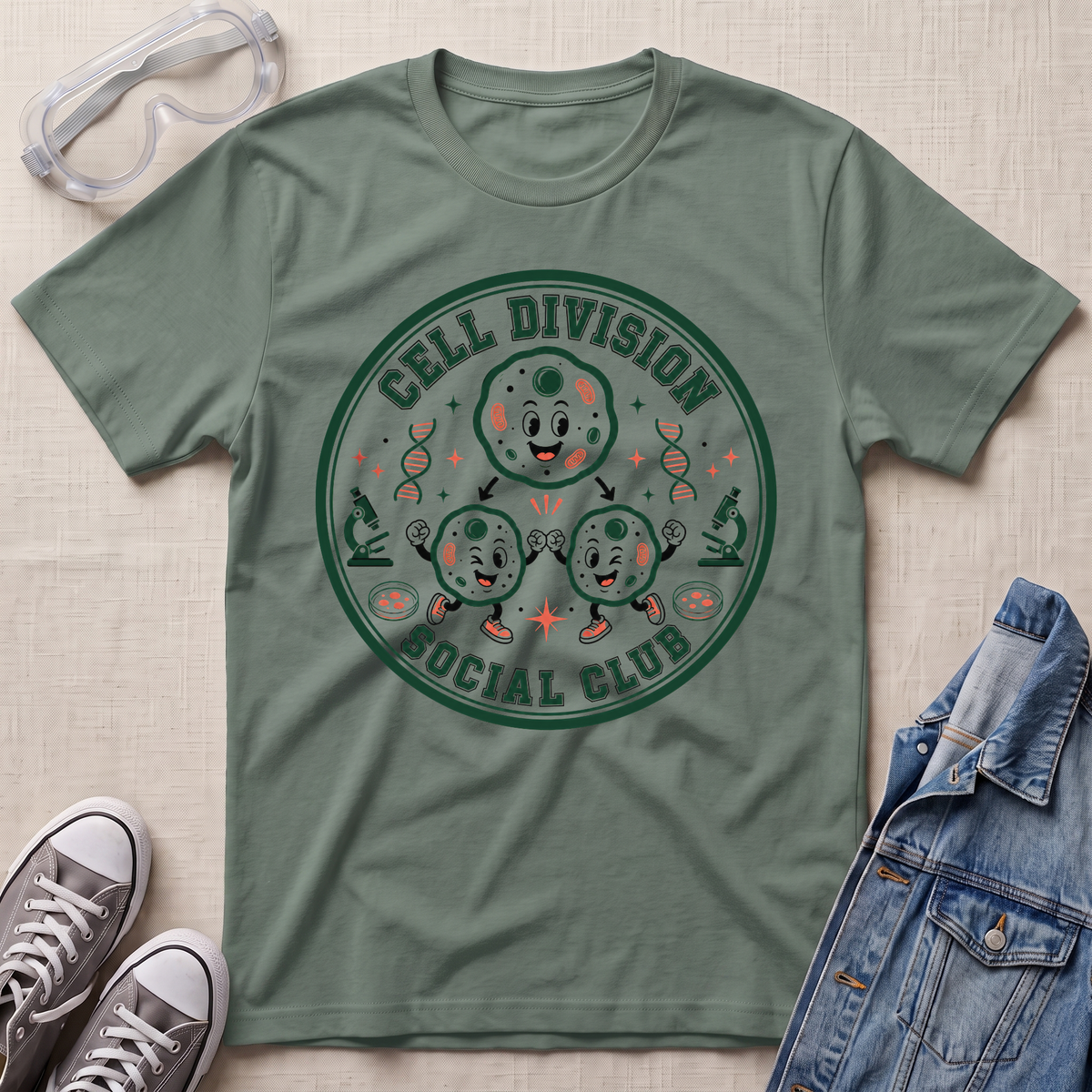 A sage green graphic t-shirt featuring a circular 'Cell Division Social Club' design with cartoon cells and microscopes, styled in a flat lay with safety goggles, sneakers, and a denim jacket on a textured beige background.