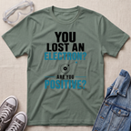 A sage green graphic t-shirt laid flat on a textured beige background, styled with safety goggles, gray sneakers, and a denim jacket. The t-shirt features a black and blue text graphic that reads 'YOU LOST AN ELECTRON? ARE YOU POSITIVE?' with a simple electron illustration.