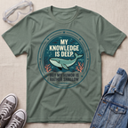 A sage green graphic t-shirt laid flat on a textured beige fabric background, styled with safety goggles, gray sneakers, and a denim jacket. The t-shirt features a circular graphic with a teal whale and the text 'MY KNOWLEDGE IS DEEP BUT MY HUMOR IS RATHER SHALLOW'.