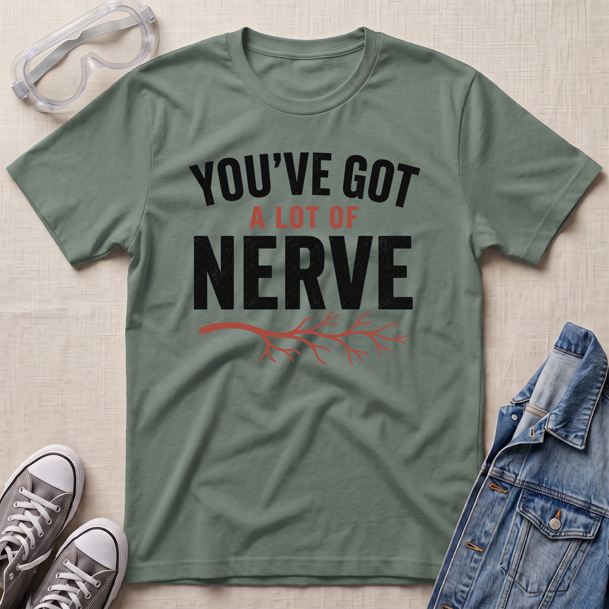 An olive green graphic t-shirt laid flat on a textured beige fabric background, surrounded by lifestyle props including clear safety goggles, gray sneakers, and a blue denim jacket. The t-shirt features the text 'YOU'VE GOT A LOT OF NERVE' in black and orange-red lettering with a red branch graphic.