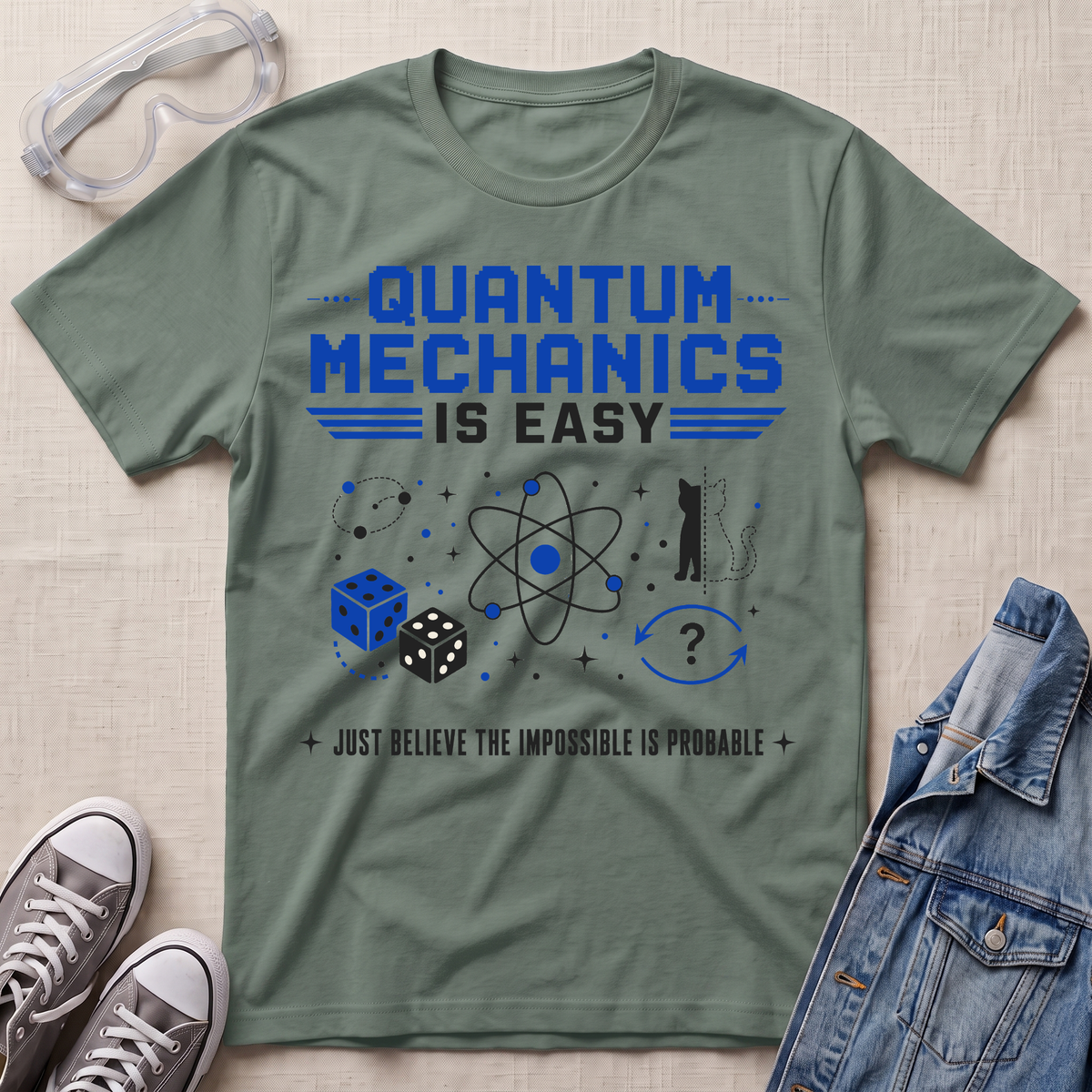 An olive green graphic t-shirt featuring blue and black text that reads 'QUANTUM MECHANICS IS EASY' with an atom and dice illustration. The t-shirt is styled in a flat-lay arrangement with safety goggles, gray sneakers, and a denim jacket on a textured beige fabric background.