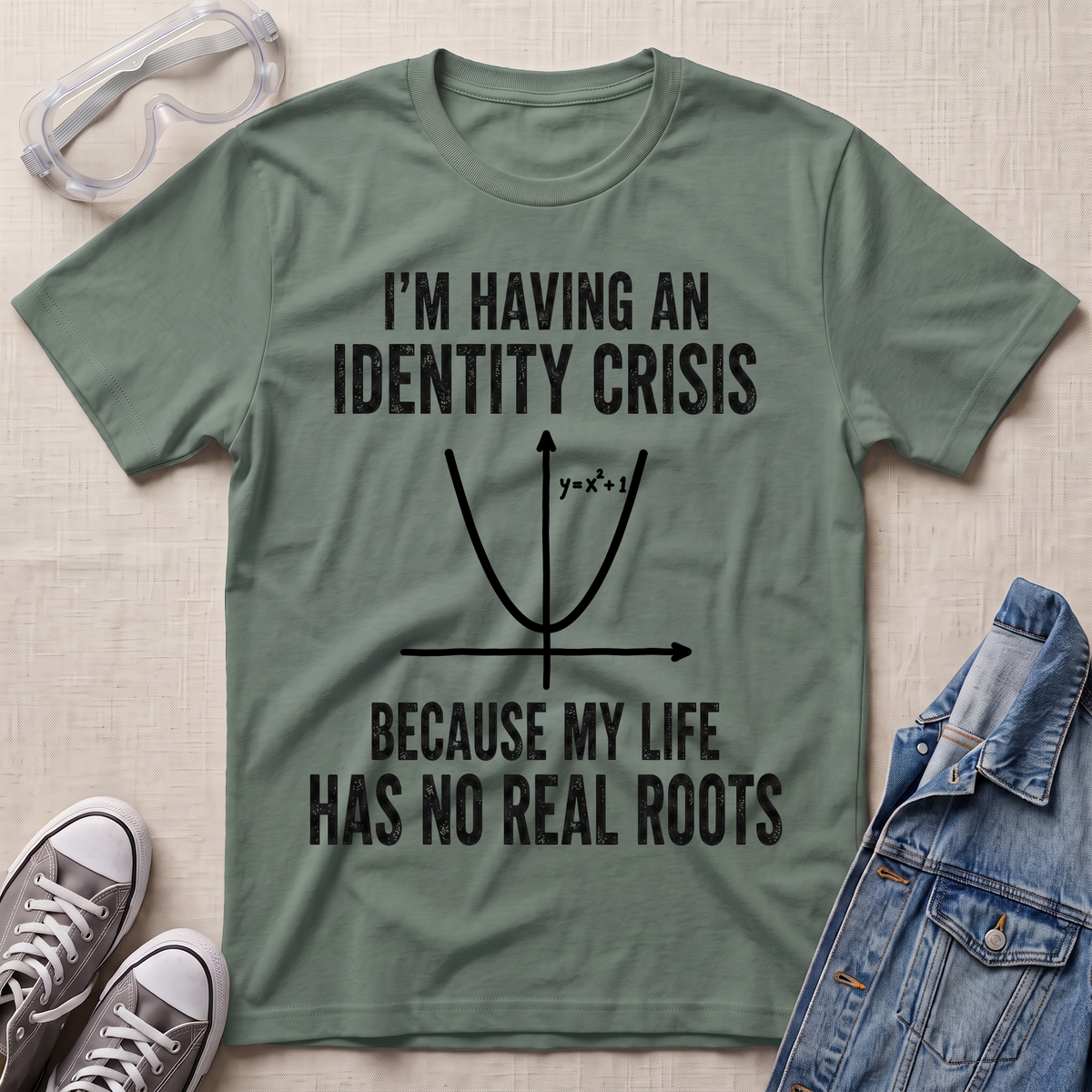 An olive green graphic t-shirt laid flat on a light beige fabric background, styled with a pair of clear safety glasses, gray sneakers, and a blue denim jacket. The t-shirt features a black printed graphic of a parabola and the text 'I'M HAVING AN IDENTITY CRISIS BECAUSE MY LIFE HAS NO REAL ROOTS'.