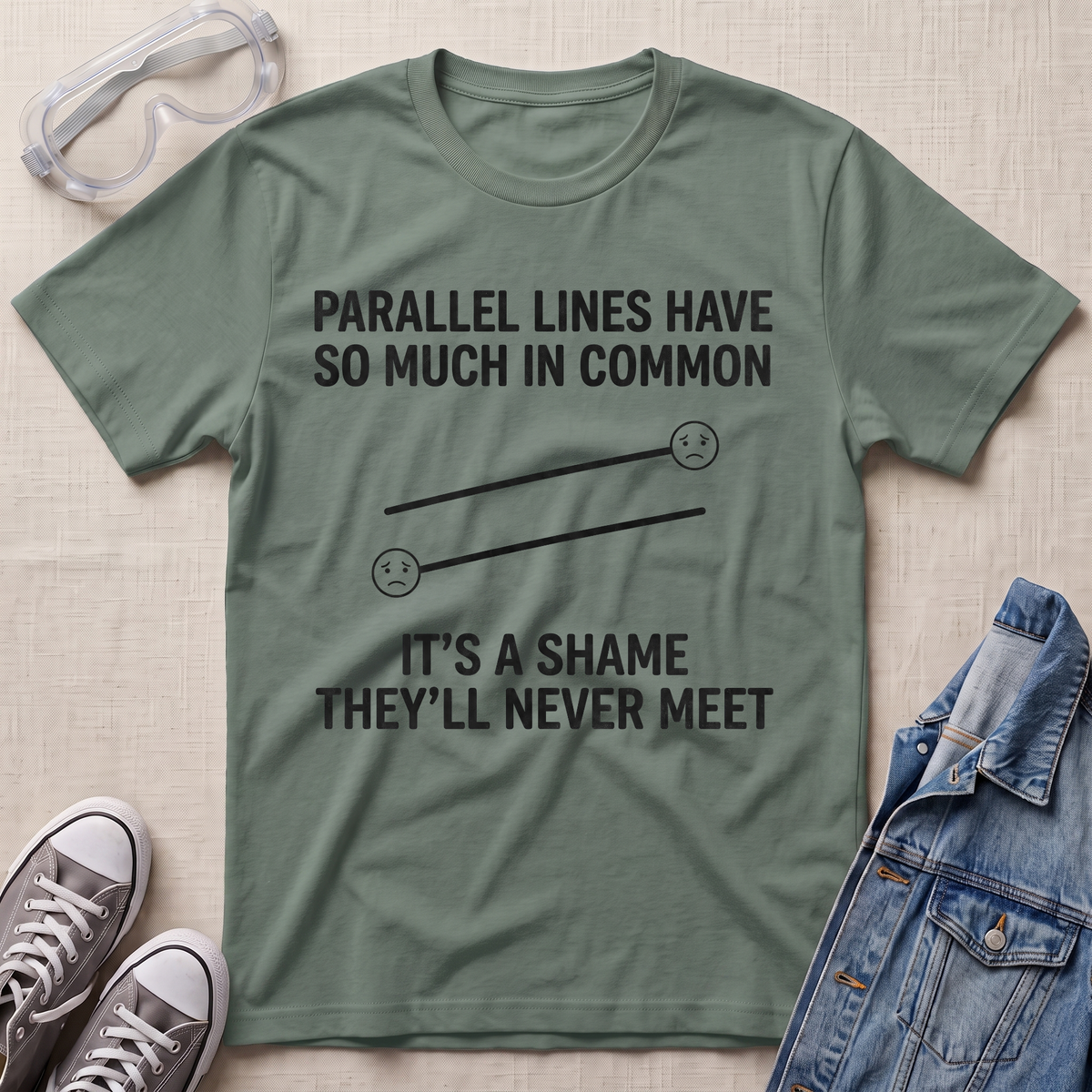 An olive green graphic t-shirt laid flat on a textured beige fabric background, surrounded by lifestyle props including safety goggles, gray sneakers, and a blue denim jacket. The t-shirt features a black printed graphic of parallel lines with sad faces and the text 'PARALLEL LINES HAVE SO MUCH IN COMMON IT'S A SHAME THEY'LL NEVER MEET'.