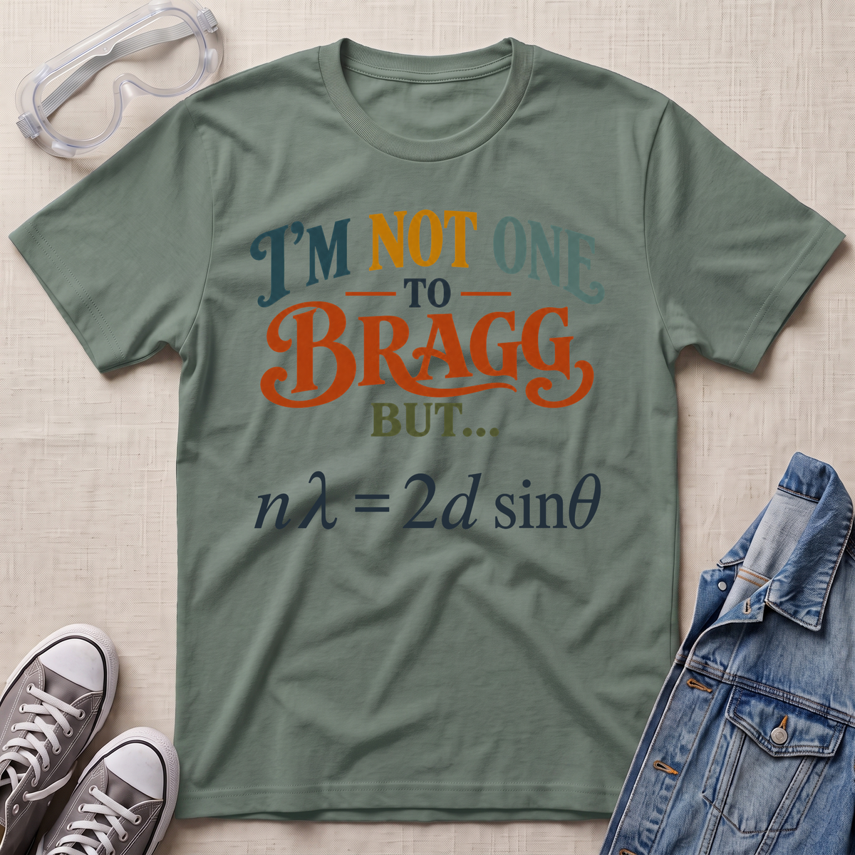 An olive green graphic t-shirt laid flat on a textured beige fabric background, surrounded by styling props including safety goggles, gray sneakers, and a blue denim jacket. The t-shirt features a humorous text and physics equation graphic.