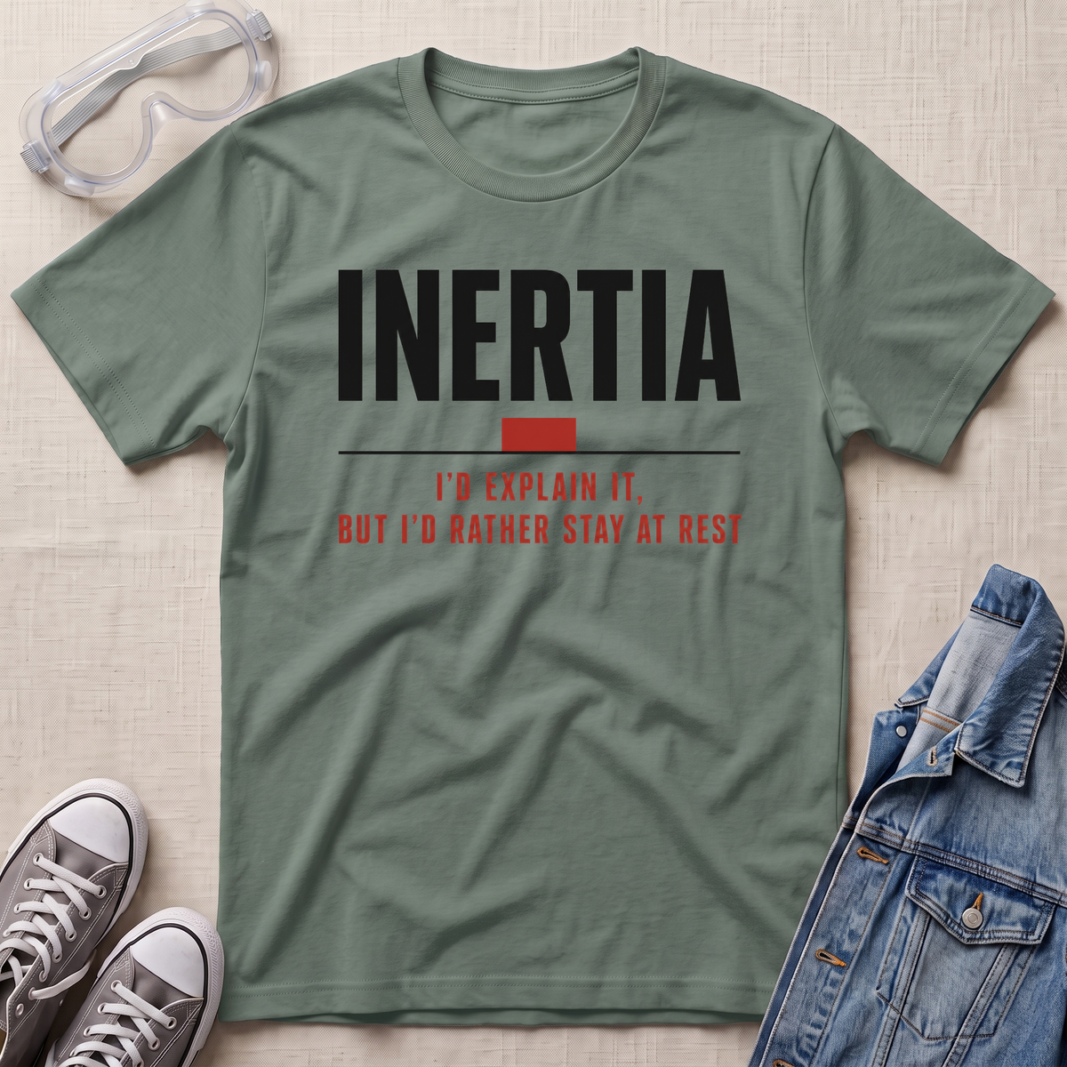 An olive green graphic t-shirt laid flat on a textured beige fabric background, surrounded by lifestyle props including clear safety goggles, gray sneakers, and a blue denim jacket. The t-shirt features the word 'INERTIA' in bold black letters with a small red block and red text underneath reading 'I'D EXPLAIN IT, BUT I'D RATHER STAY AT REST'.