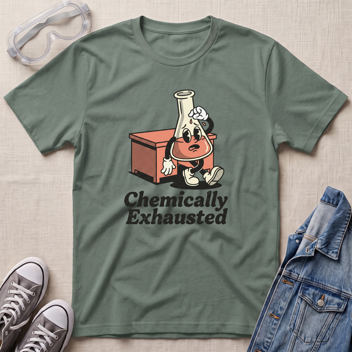 An olive green graphic t-shirt laid flat on a textured beige background, surrounded by lifestyle props including safety goggles, gray sneakers, and a denim jacket. The t-shirt features a cartoon graphic of a chemist character with an Erlenmeyer flask on their head, sitting next to an orange beaker, with the text 'Chemically Exhausted' printed below.