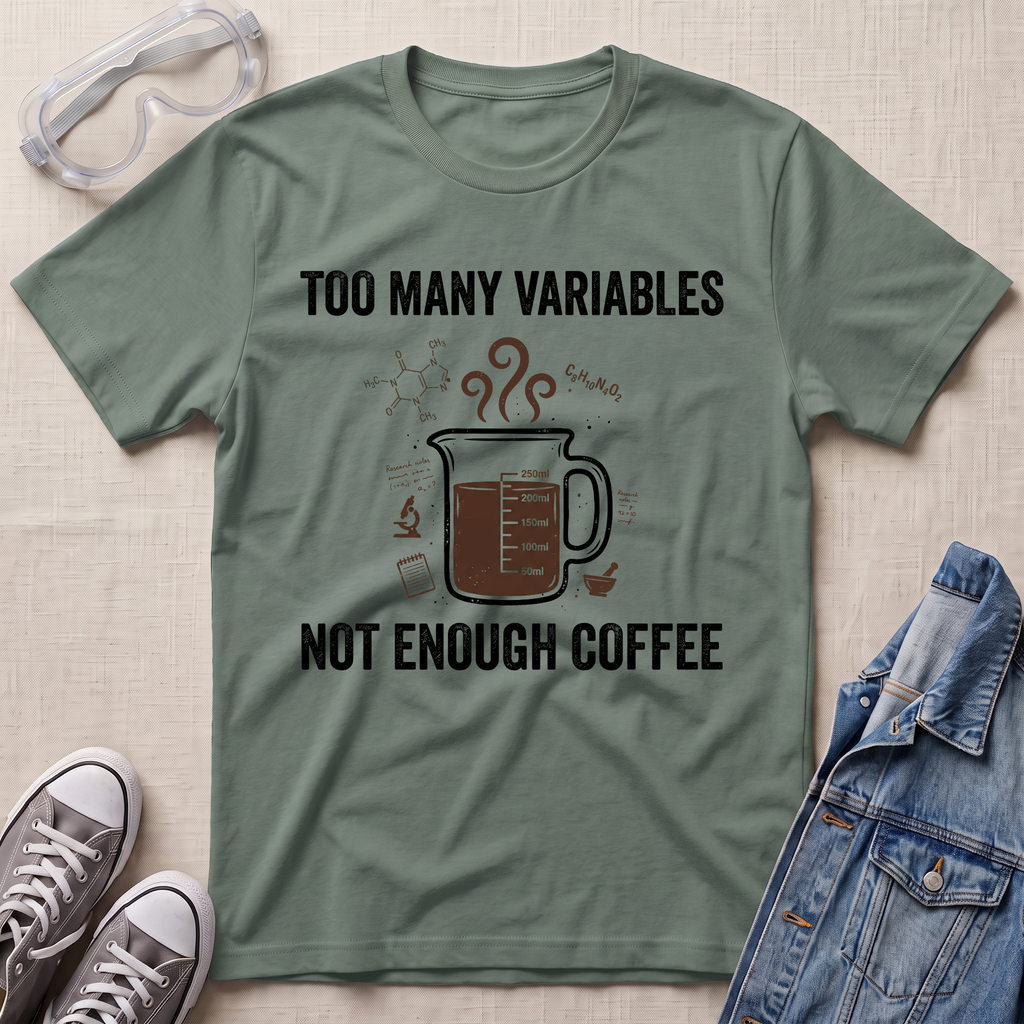 An olive green graphic t-shirt laid flat on a textured beige fabric background, styled with safety goggles, gray sneakers, and a denim jacket. The t-shirt features a dark brown coffee mug graphic with steam and the text 'TOO MANY VARIABLES NOT ENOUGH COFFEE' in black.