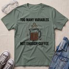 An olive green graphic t-shirt laid flat on a textured beige fabric background, styled with safety goggles, gray sneakers, and a denim jacket. The t-shirt features a dark brown coffee mug graphic with steam and the text 'TOO MANY VARIABLES NOT ENOUGH COFFEE' in black.