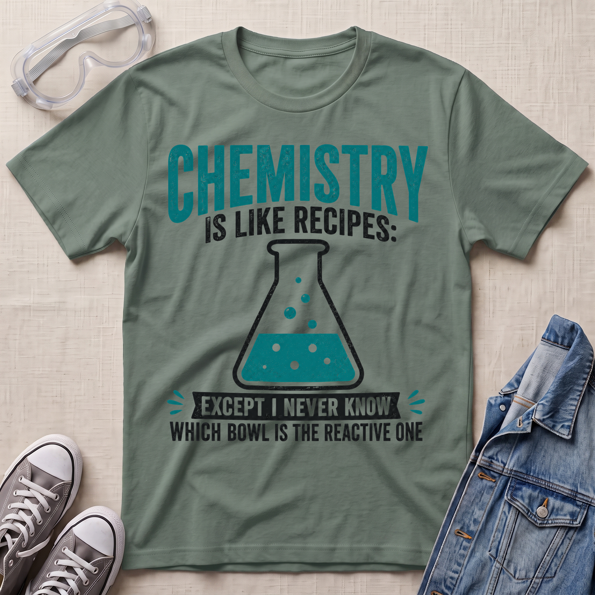 An olive green graphic t-shirt laid flat on a textured beige fabric background, styled with safety goggles, gray sneakers, and a denim jacket. The t-shirt features a teal and black graphic that reads 'CHEMISTRY IS LIKE RECIPES: EXCEPT I NEVER KNOW WHICH BOWL IS THE REACTIVE ONE' with an illustration of a beaker.