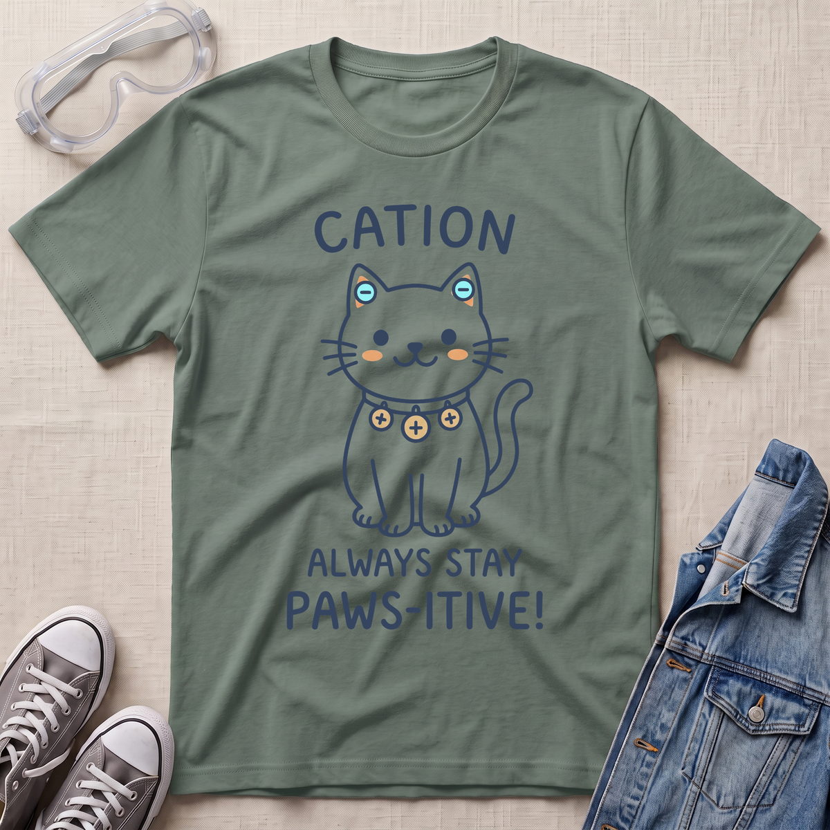 An olive green graphic t-shirt laid flat on a textured beige fabric background, styled with safety goggles, gray sneakers, and a denim jacket. The t-shirt features a cartoon cat wearing a heart-patterned collar with the text 'CATION ALWAYS STAY PAWS-ITIVE!'.