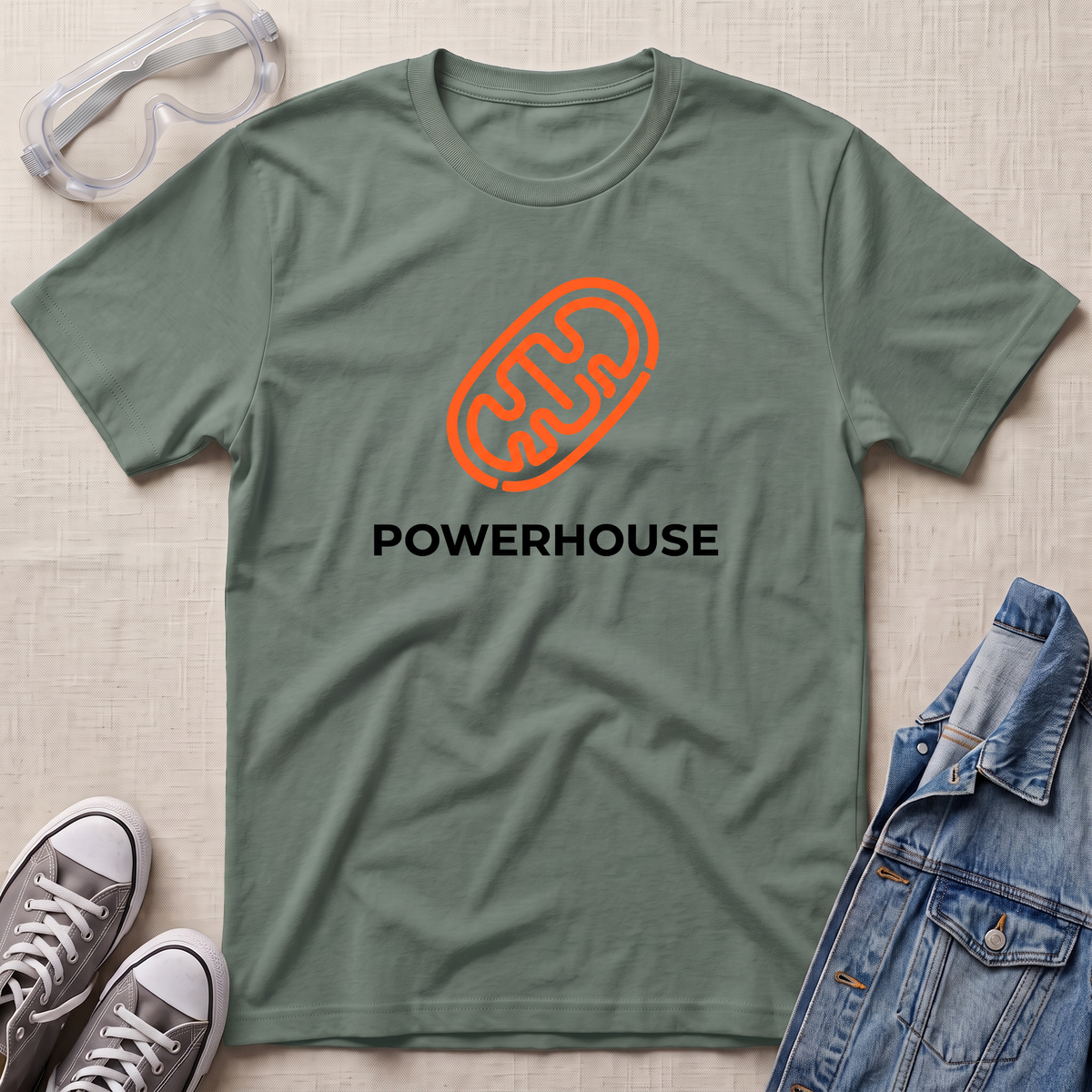 An olive green crewneck t-shirt with an orange mitochondrion outline and the word 'POWERHOUSE' printed in black, styled in a flat lay with safety goggles, gray sneakers, and a denim jacket on a beige textured background.