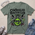 A sage green graphic t-shirt featuring a cartoon beaker with sunglasses and the text 'CONTROLLED REACTION CLUB' in black. The shirt is styled in a flat-lay arrangement with safety goggles, gray sneakers, and a denim jacket on a textured beige background.