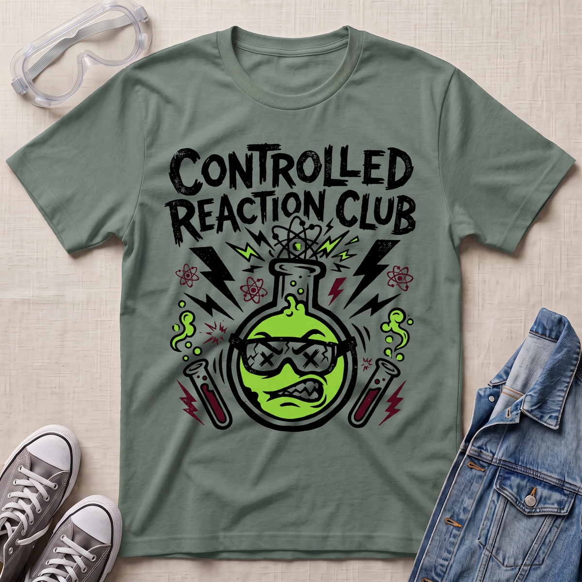 A sage green graphic t-shirt featuring a cartoon beaker with sunglasses and the text 'CONTROLLED REACTION CLUB' in black. The shirt is styled in a flat-lay arrangement with safety goggles, gray sneakers, and a denim jacket on a textured beige background.