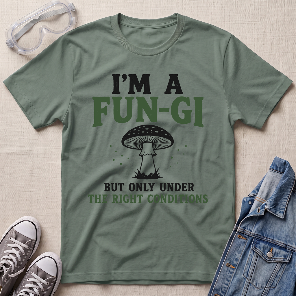 A sage green graphic t-shirt laid flat on a textured beige fabric background, styled with safety glasses, gray sneakers, and a blue denim jacket. The t-shirt features a black and dark green graphic of a mushroom with the text 'I'M A FUN-GI BUT ONLY UNDER THE RIGHT CONDITIONS'.