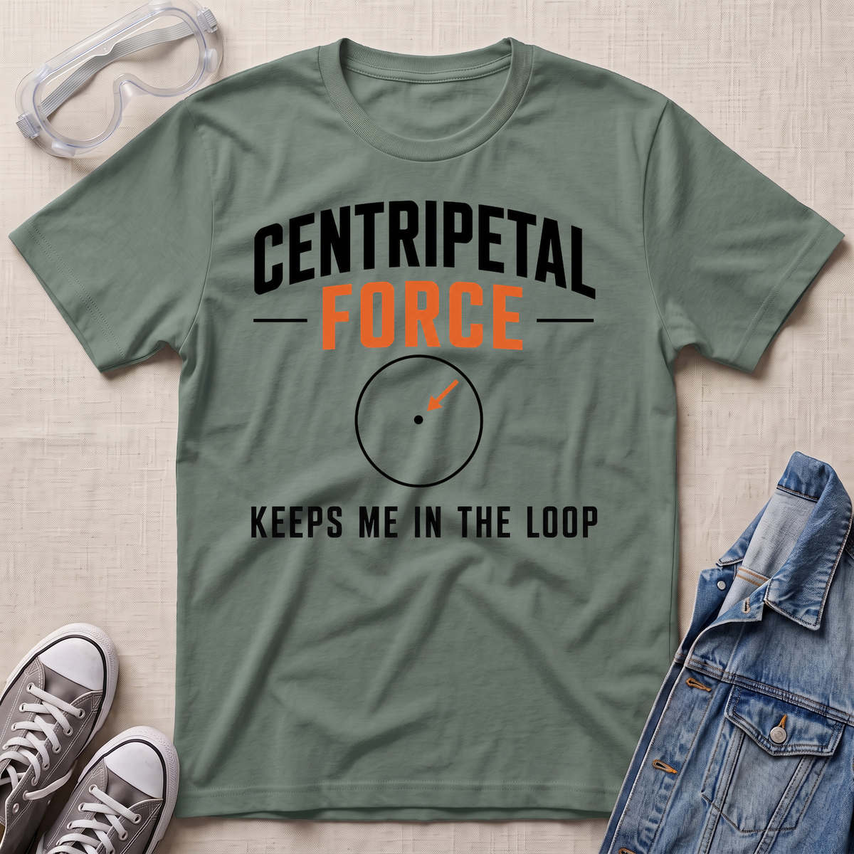 An olive green graphic t-shirt laid flat on a beige textured background, surrounded by styling props including safety goggles, gray sneakers, and a blue denim jacket. The t-shirt features the text 'CENTRIPETAL FORCE' in black and orange, with a circular diagram graphic below it and the phrase 'KEEPS ME IN THE LOOP'.