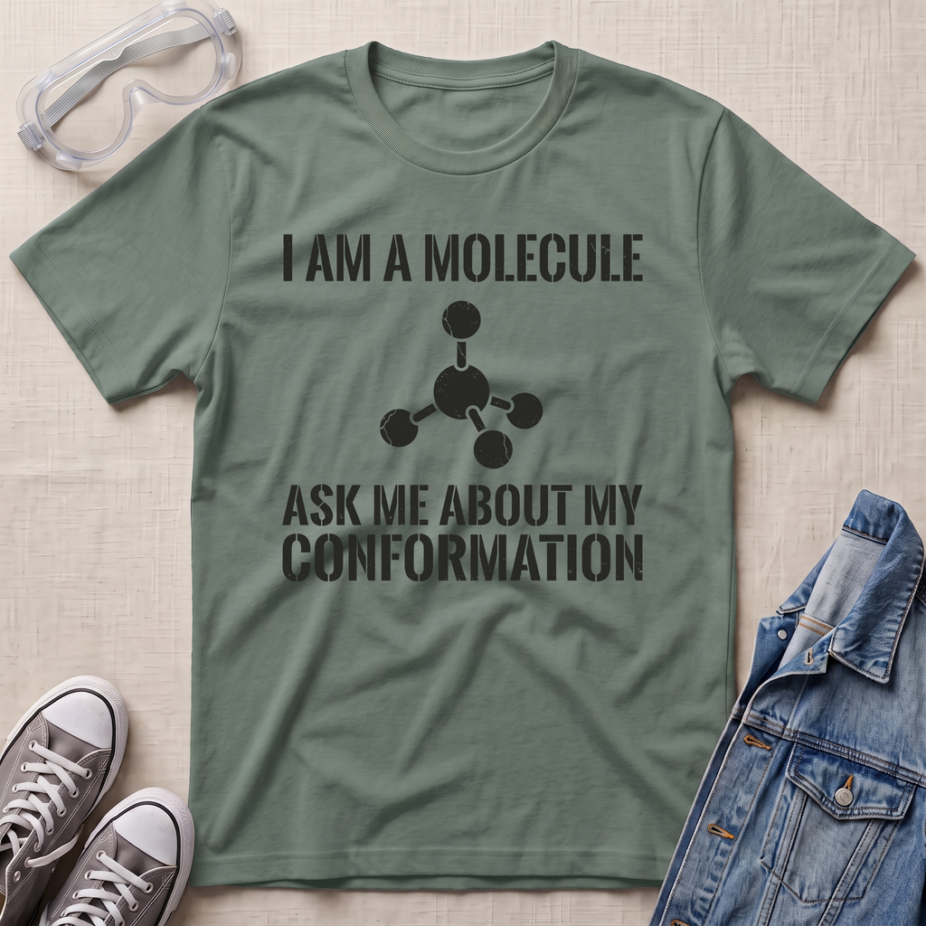 An olive green graphic t-shirt laid flat on a light beige textured background, styled with safety goggles, gray sneakers, and a blue denim jacket. The t-shirt features a black printed graphic of a molecule and the text 'I AM A MOLECULE ASK ME ABOUT MY CONFORMATION'.