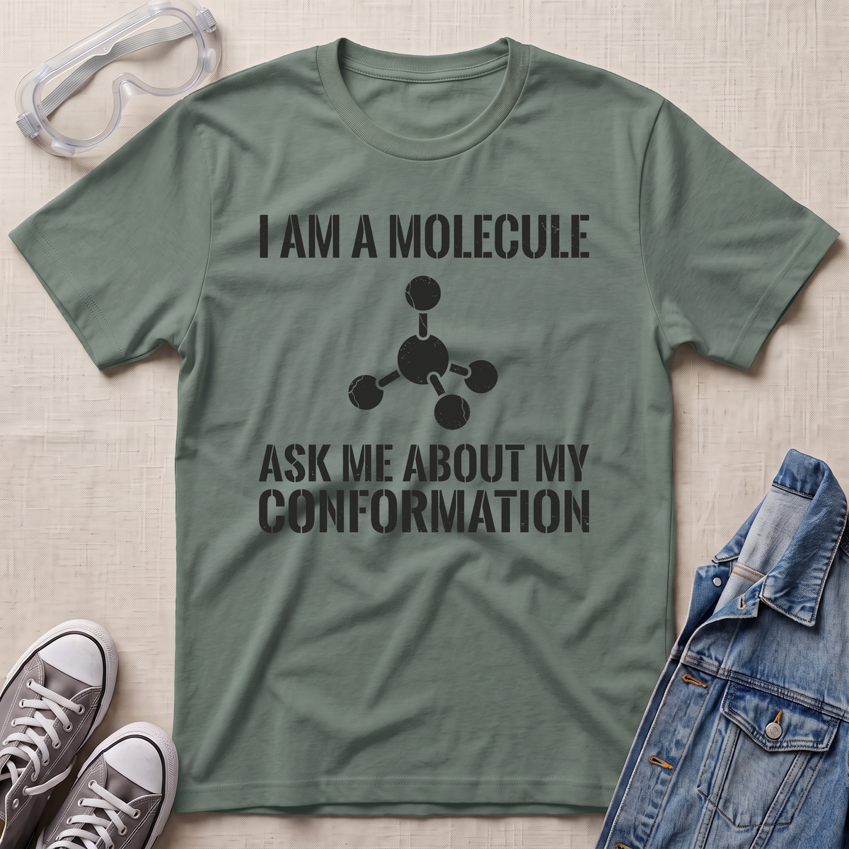 An olive green graphic t-shirt laid flat on a light beige textured background, styled with safety goggles, gray sneakers, and a blue denim jacket. The t-shirt features a black printed graphic of a molecule and the text 'I AM A MOLECULE ASK ME ABOUT MY CONFORMATION'.