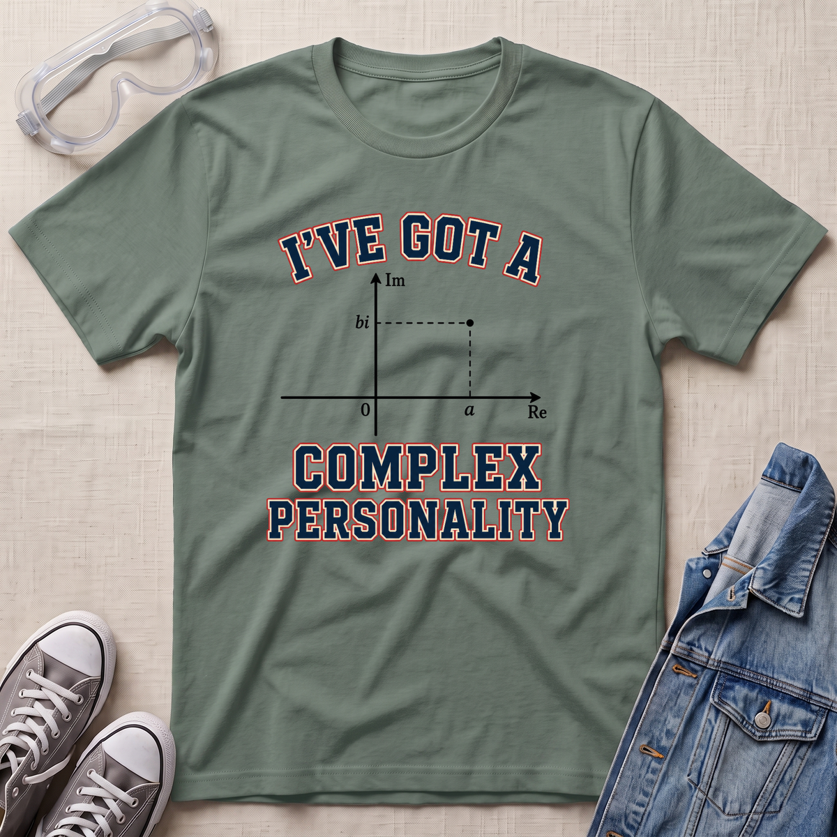 An olive green graphic t-shirt laid flat on a textured beige fabric background, styled with safety goggles, gray sneakers, and a denim jacket. The t-shirt features a physics-themed graphic with a frequency graph and the text 'I'VE GOT A COMPLEX PERSONALITY'.