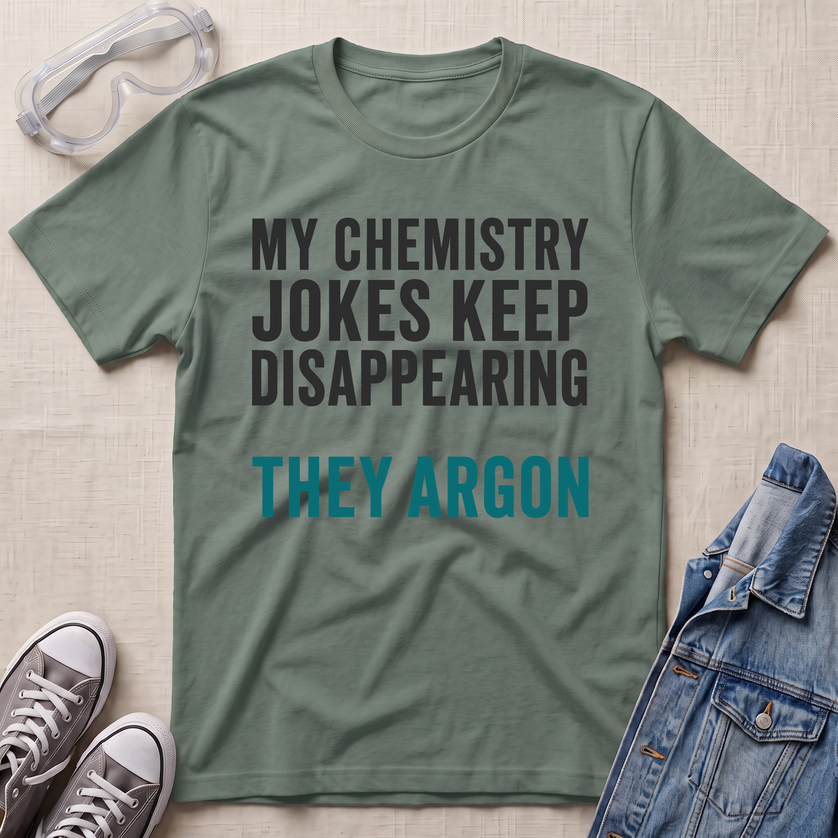 An olive green graphic t-shirt laid flat on a textured beige fabric background, surrounded by styling props including safety goggles, gray sneakers, and a blue denim jacket. The t-shirt features the text 'MY CHEMISTRY JOKES KEEP DISAPPEARING' in dark gray, and 'THEY ARGON' in teal.