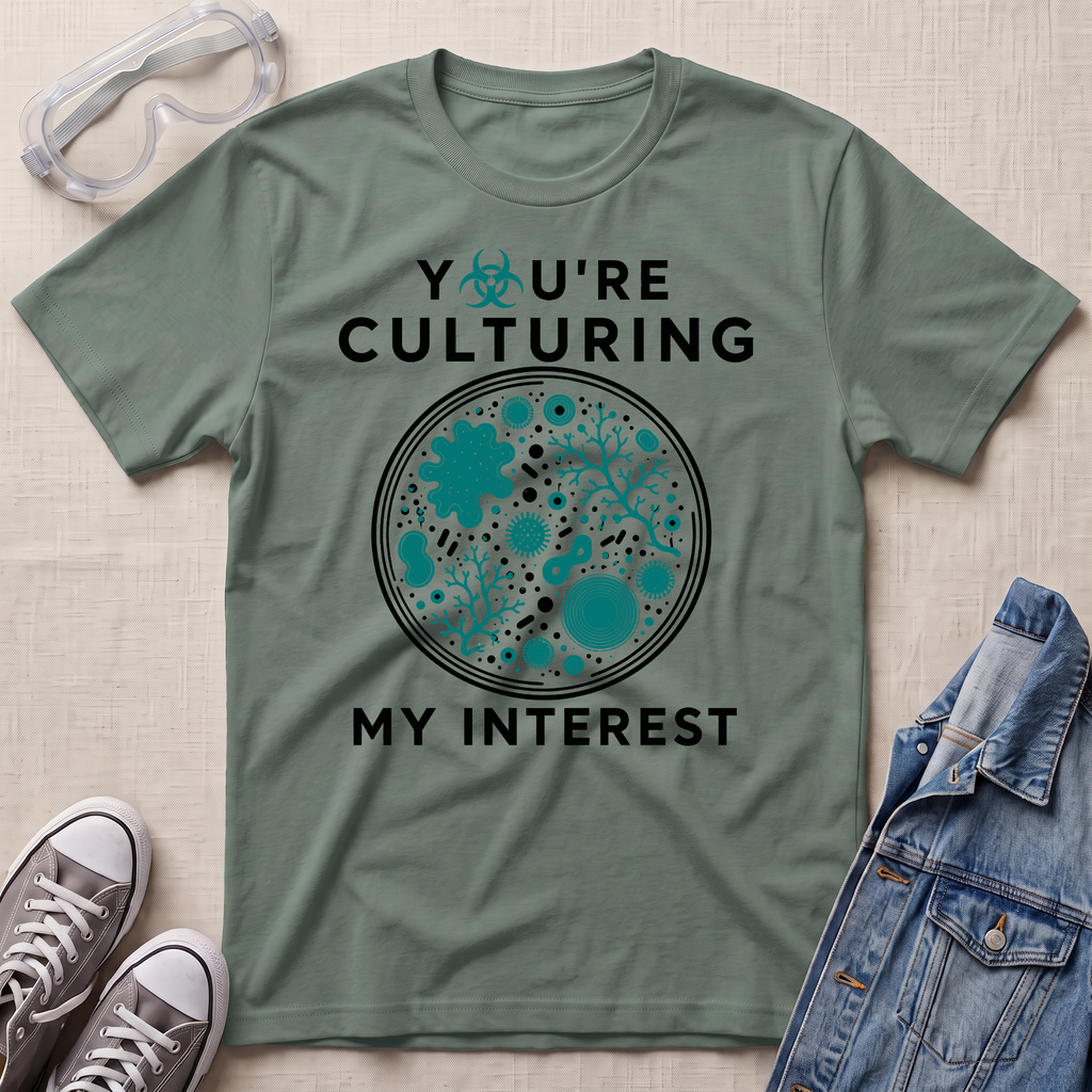 An olive green graphic t-shirt laid flat on a light beige textured background, surrounded by props including safety goggles, gray sneakers, and a blue denim jacket. The t-shirt features a circular petri-dish style graphic with teal and black microorganisms and the text 'YOU'RE CULTURING MY INTEREST' in black and teal.