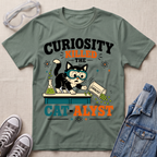 A sage green graphic t-shirt laid flat on a textured beige fabric background, styled with safety goggles, gray sneakers, and a denim jacket. The t-shirt features a cartoon of a curious black and white cat wearing goggles and looking at a beaker, with the text 'CURIOSITY KILLED THE CAT-ALYST'.