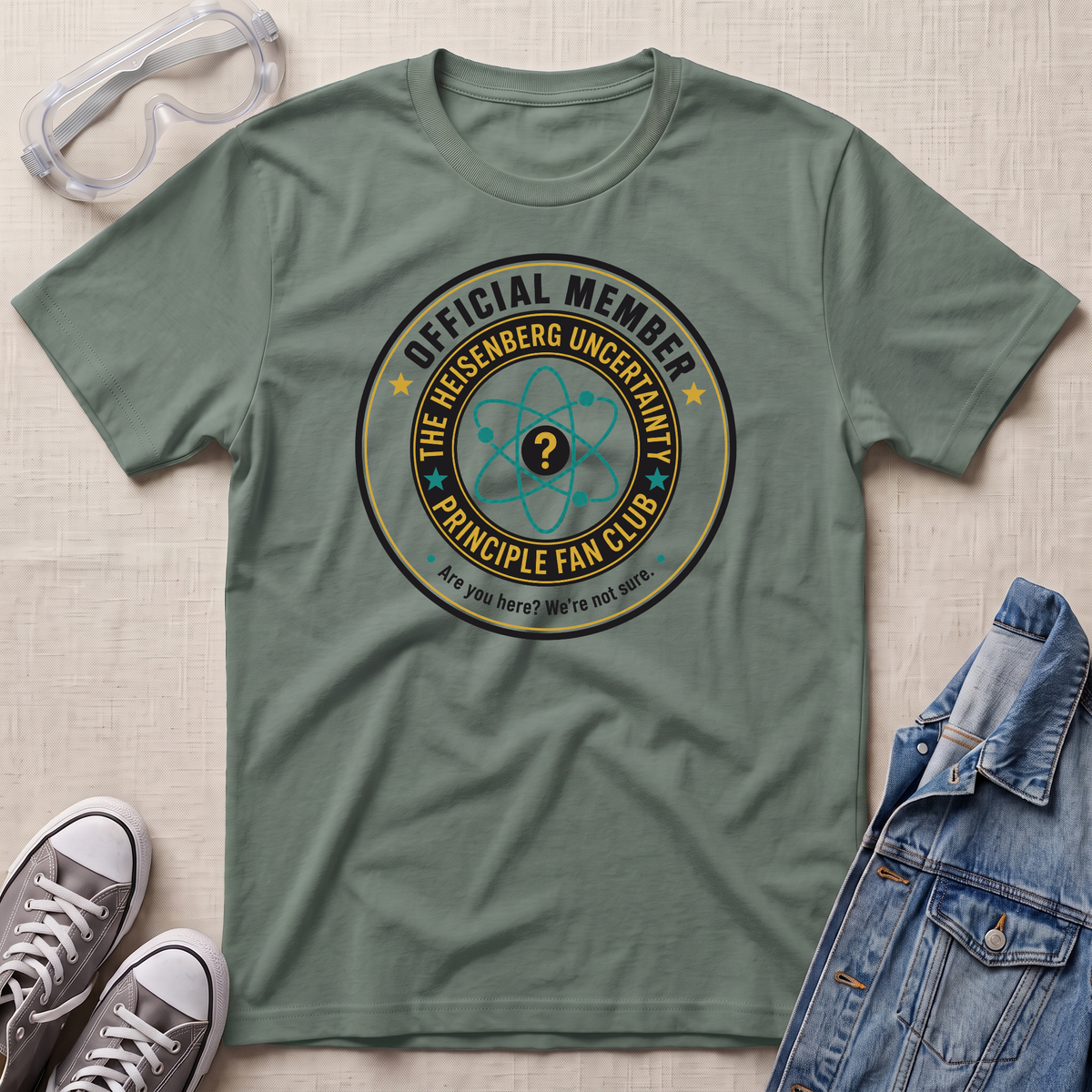 An olive green graphic t-shirt laid flat on a textured beige fabric background, styled with clear safety goggles, gray sneakers, and a blue denim jacket. The t-shirt features a circular graphic that reads 'OFFICIAL MEMBER THE HEISENBERG UNCERTAINTY PRINCIPLE FAN CLUB' with an atom symbol.