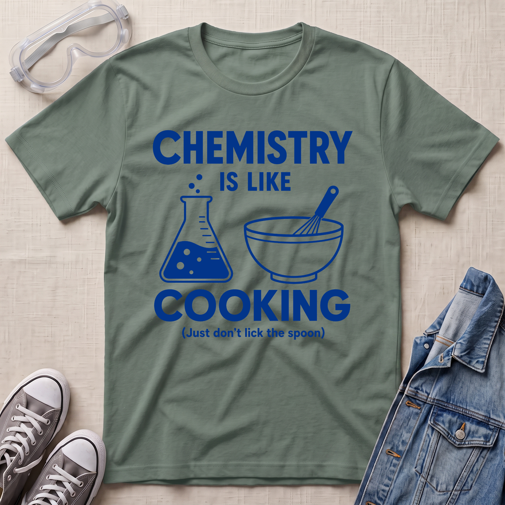 An olive green graphic t-shirt laid flat on a textured beige background, surrounded by styling props including safety goggles, sneakers, and a denim jacket. The t-shirt features a blue graphic that reads 'CHEMISTRY IS LIKE COOKING (Just don't lick the spoon)' with illustrations of a beaker and a bowl with a whisk.