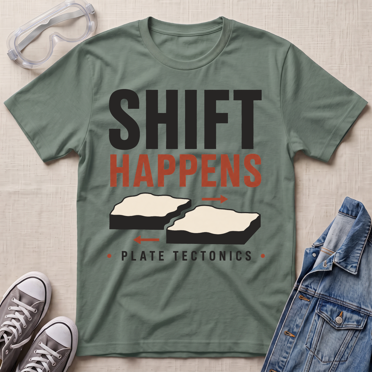 An olive green graphic t-shirt laid flat on a textured beige background, styled with safety goggles, gray sneakers, and a denim jacket. The t-shirt features a graphic that reads 'SHIFT HAPPENS' in black and orange, with illustrations of splitting landmasses and 'PLATE TECTONICS' underneath.