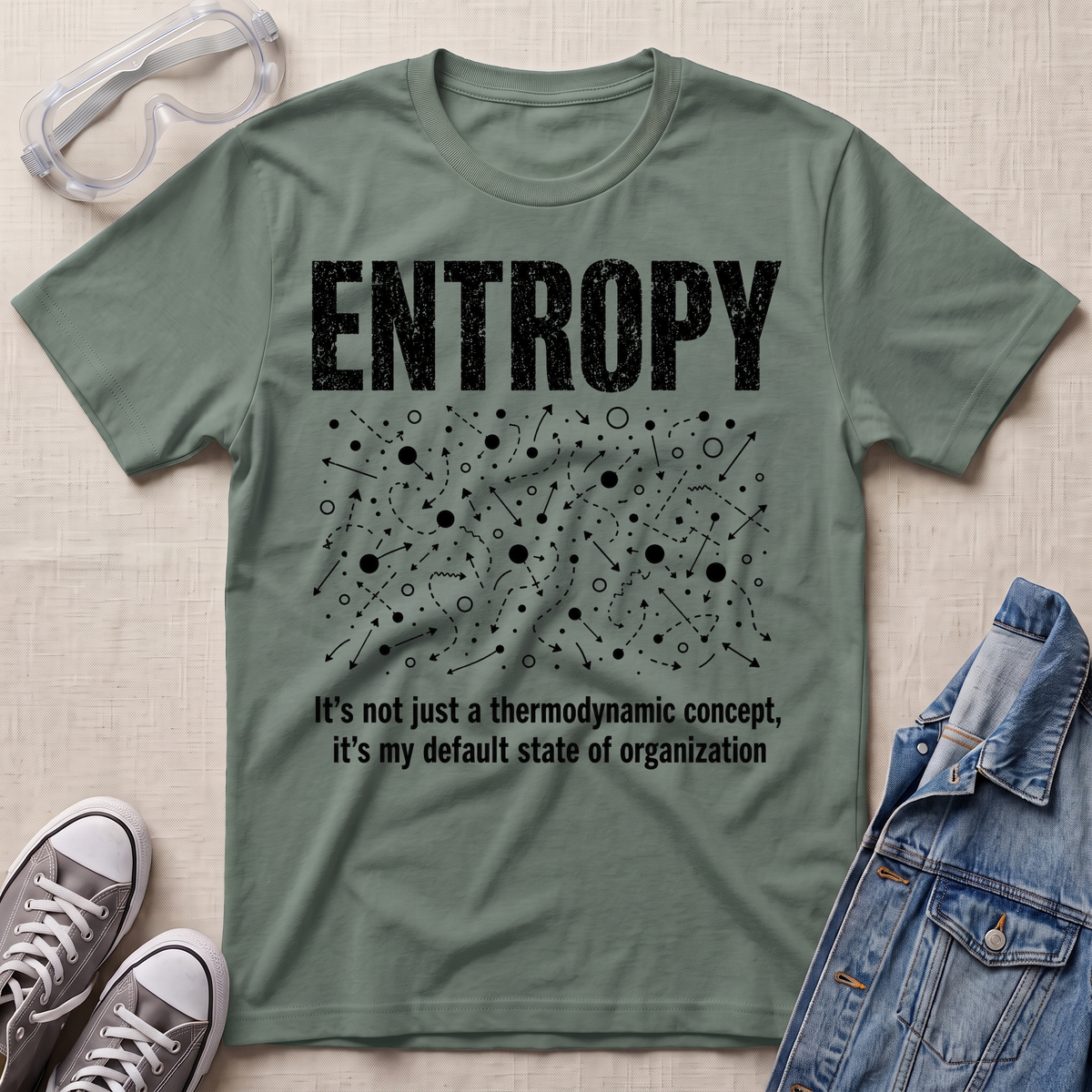 A sage green graphic t-shirt laid flat on a beige fabric background, styled with safety goggles, gray sneakers, and a denim jacket. The t-shirt features a white distressed graphic of the word 'ENTROPY' above a entropy pattern, with the text 'It's not just a thermodynamic concept, it's my default state of organization' below.