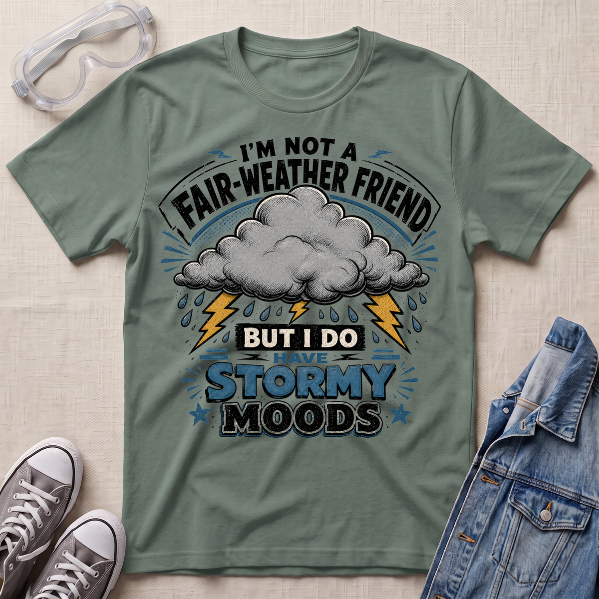 An olive green graphic t-shirt laid flat on a textured beige background, styled with clear safety goggles, gray sneakers, and a blue denim jacket. The t-shirt features a storm graphic with a gray cloud, yellow lightning bolts, and blue text that reads 'I'M NOT A FAIR-WEATHER FRIEND BUT I DO HAVE STORMY MOODS'.