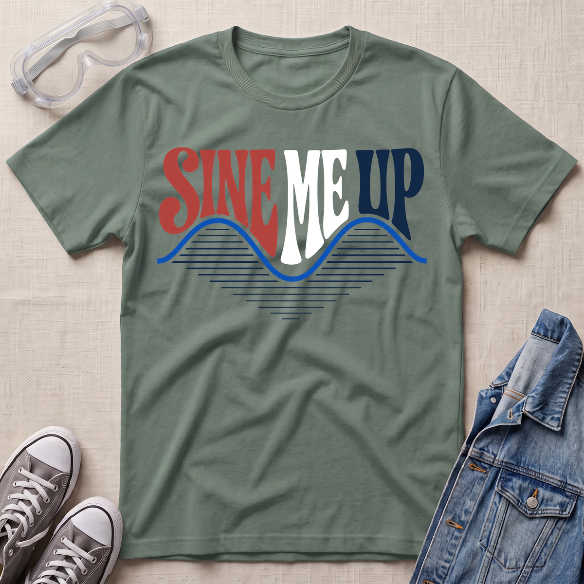 An olive green graphic t-shirt laid flat on a textured beige fabric background, surrounded by styling props including clear safety goggles, gray sneakers, a denim jacket, and a pair of jeans. The t-shirt features a retro-style graphic reading 'SINE ME UP' in red, white, and blue, with a wavy blue line underneath.