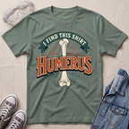 A sage green graphic t-shirt laid flat on a light wood surface, featuring a bone graphic and the text 'I FIND THIS SHIRT HUMERUS' in orange and green. The shirt is styled with safety glasses, gray sneakers, and a denim jacket.