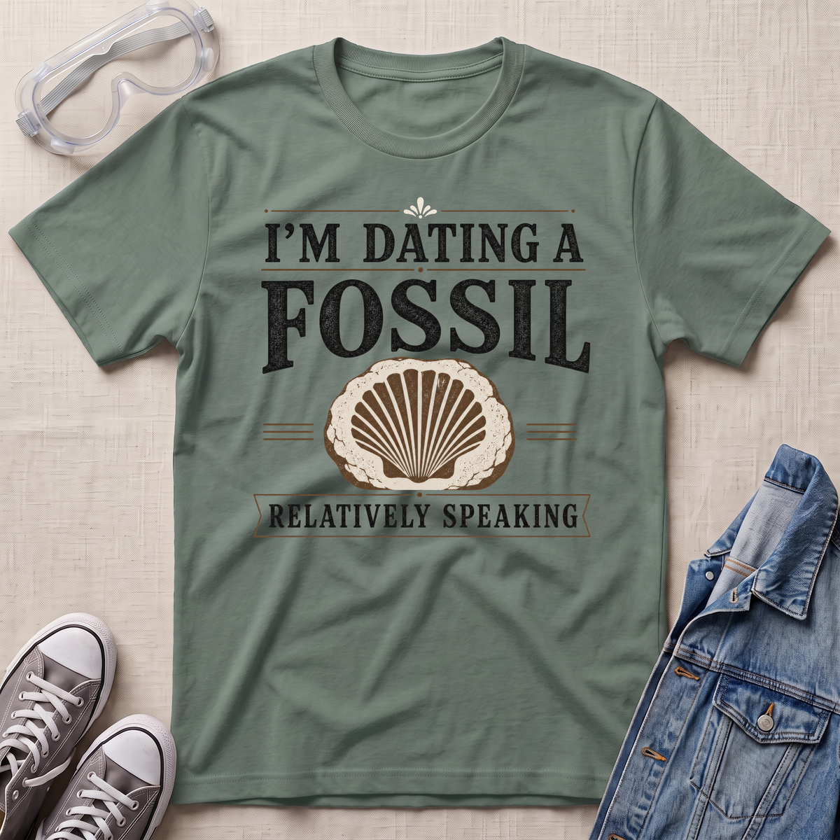 An olive green graphic t-shirt laid flat on a textured beige background, styled with safety goggles, gray sneakers, and a denim jacket. The t-shirt features a distressed black and beige graphic that reads 'I'M DATING A FOSSIL RELATIVELY SPEAKING' with a seashell illustration.