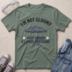 An olive green graphic t-shirt laid flat on a textured beige background, styled with safety goggles, gray sneakers, and a blue denim jacket. The t-shirt features a dark gray cloud graphic and the text 'I'M NOT GLOOMY JUST UNDER A LITTLE PRESSURE' with two clock illustrations.